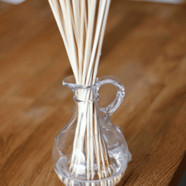 A clear glass diffuser bottle with several beige reed sticks inserted, perfect to make your own reed diffuser, placed on a wooden surface.