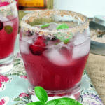 A reddish Cranberry Basil Margarita with ice cubes, garnished with berries and basil, served in a glass with a sugared rim on a patterned surface.