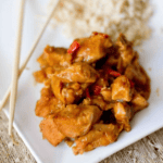 Plate of slow cooker orange chicken with red peppers and sauce served next to a portion of rice and a pair of chopsticks on a white plate.