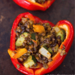 A halved red bell pepper stuffed with a mixture of lentils, diced vegetables, and herbs on a dark brown surface. These Vegan Lentil Stuffed Peppers are the perfect low carb high protein option for a nutritious meal.