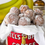 A container filled with powdered sugar-coated doughnut holes, surrounded by bananas and a bottle in the background, brings to mind the delicious treat of 10 Minute Baked Banana Bread Doughnut Holes.