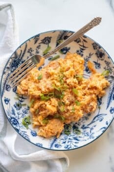 A blue and white plate with Pizza Eggs—scrambled eggs topped with chopped green onions—placed next to a folded white napkin and a fork resting on the dish.