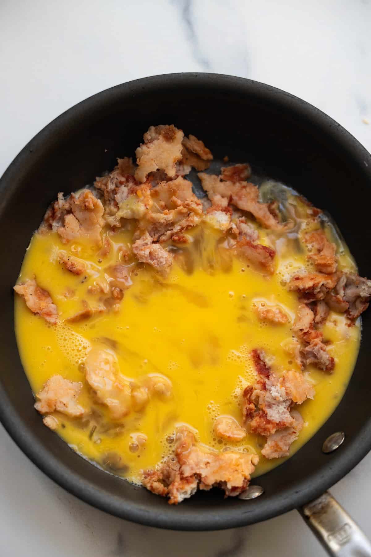 A frying pan with whisked eggs poured over pieces of cooked breaded meat, ready to be cooked on a white countertop—perfect for making savory Pizza Eggs.
