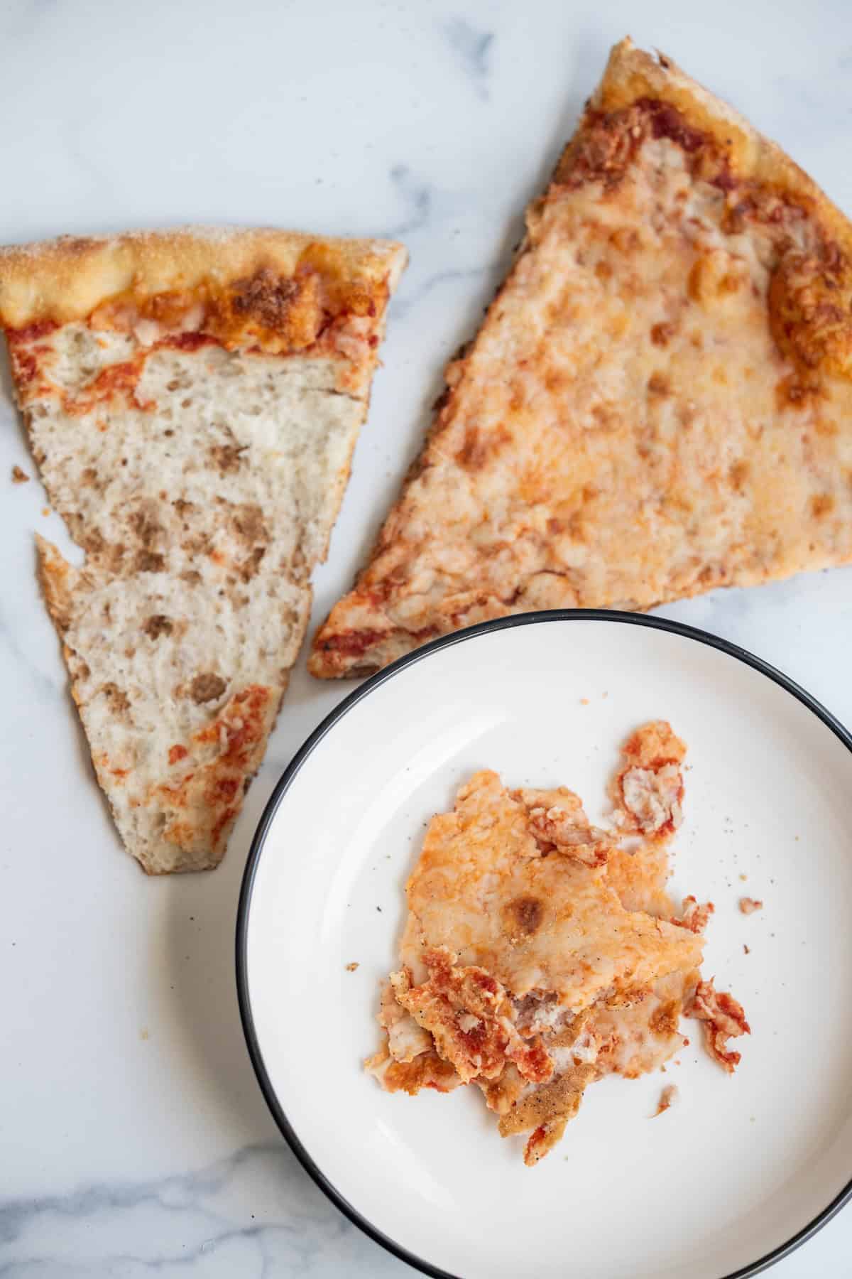 Two slices of cheese pizza, one mostly eaten, sit on a marble surface next to a plate with pizza crust and toppings scraps—perfect leftovers for making delicious Pizza Eggs the next morning.