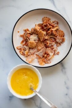 A plate with crumbled cooked food and a bowl of beaten eggs with a spoon, placed on a white marble surface—perfect for preparing delicious Pizza Eggs.