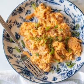 A plate of cheesy scrambled eggs, reminiscent of Pizza Eggs, garnished with chopped green onions and served in a blue and white floral-patterned bowl with a fork.