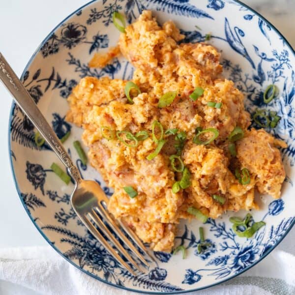 A plate of cheesy scrambled eggs, reminiscent of Pizza Eggs, garnished with chopped green onions and served in a blue and white floral-patterned bowl with a fork.