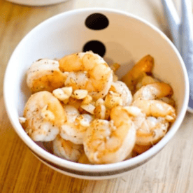 A bowl of salt and pepper shrimp garnished with garlic, served on a wooden table with utensils beside the bowl.