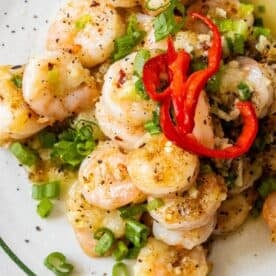 Salt and pepper shrimp cooked to perfection, garnished with sliced green onions, red chili peppers, and ground black pepper on a plate.