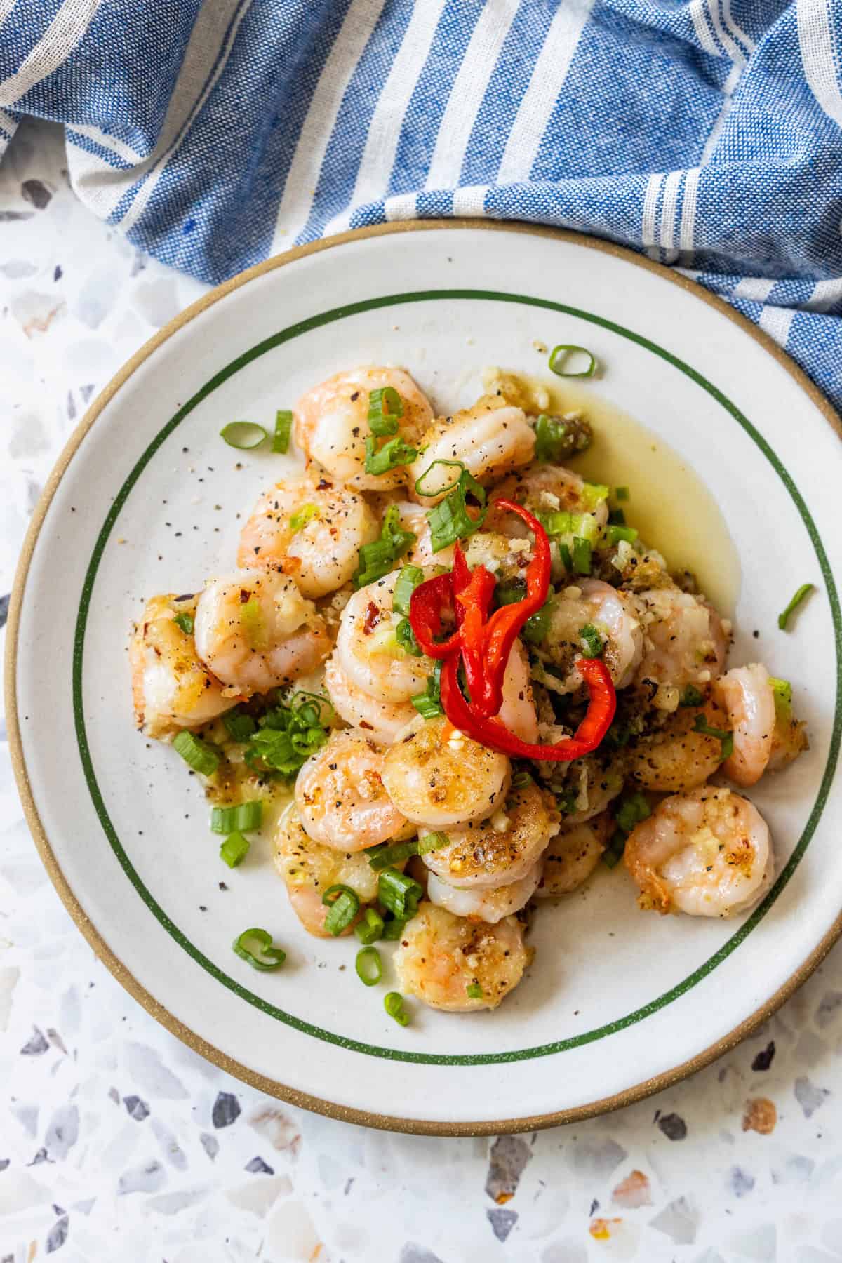 A plate of salt and pepper shrimp garnished with chopped green onions and sliced red chili peppers, set on a patterned surface beside a blue and white striped cloth.