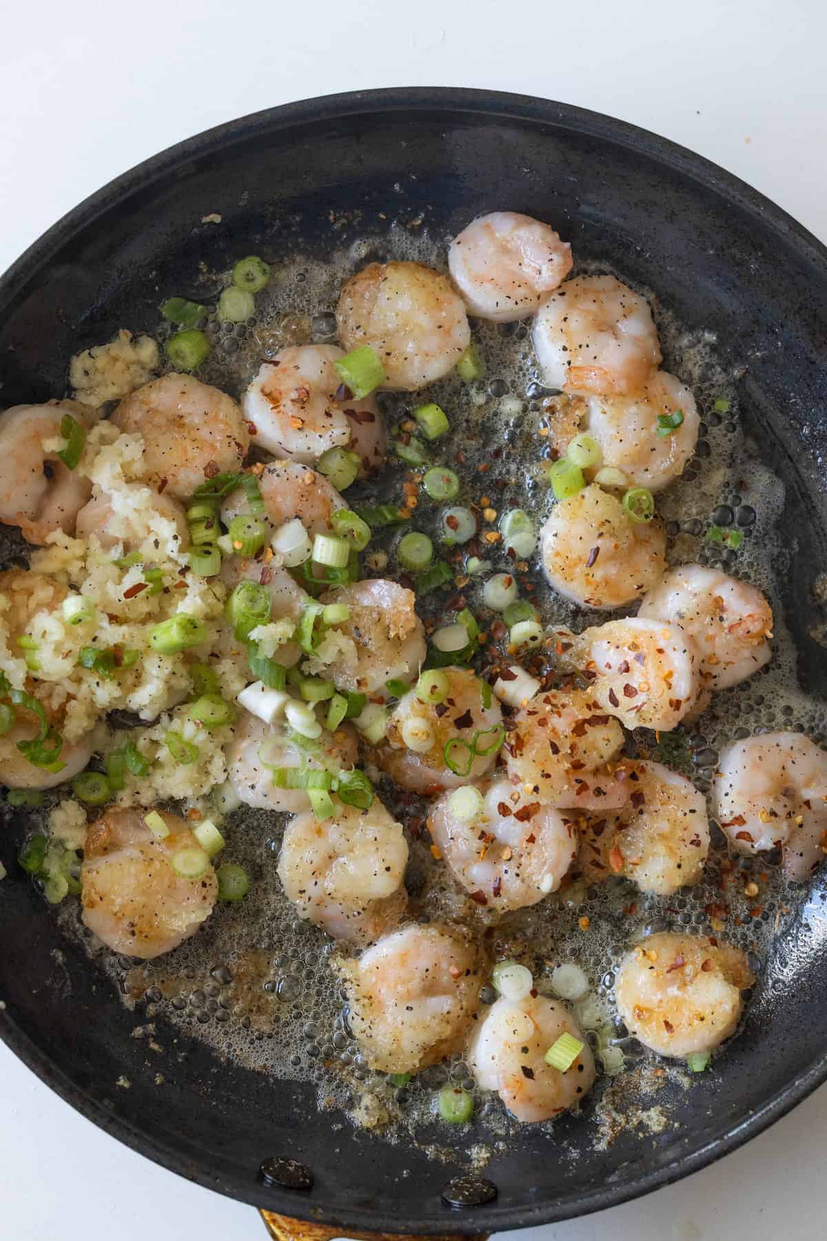Raw salt and pepper shrimp, chopped green onions, minced garlic, and red pepper flakes sizzle in a black skillet with melted butter.