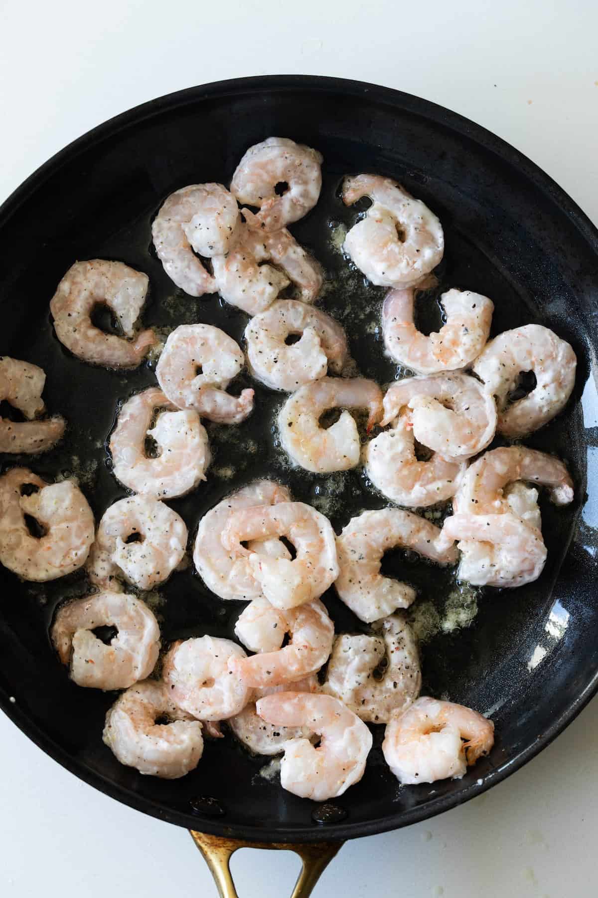 Salt and pepper shrimp being cooked in a black frying pan with melted butter on a white background.