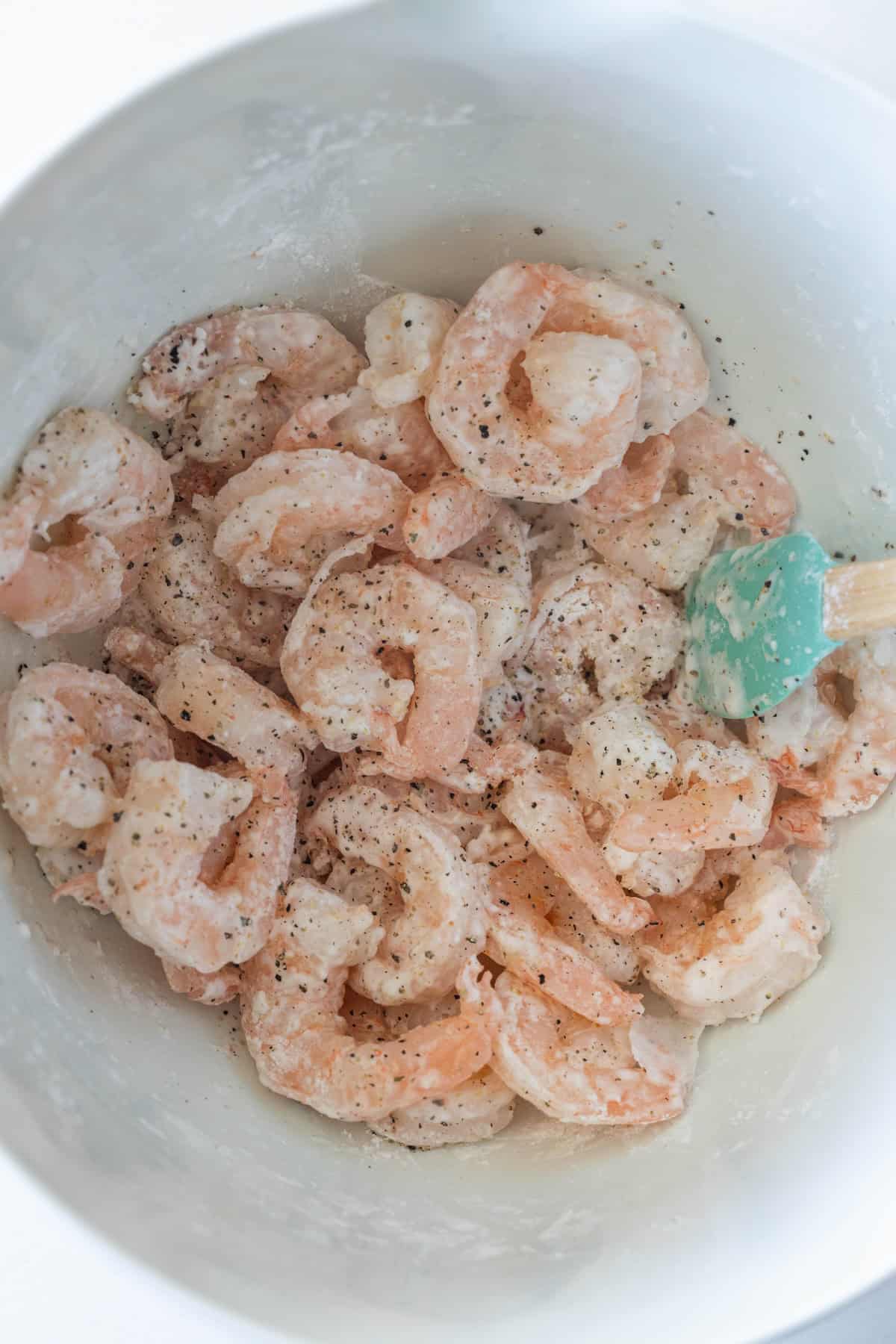 Raw shrimp for salt and pepper shrimp coated with seasoning and flour in a white bowl, being mixed with a teal spatula.