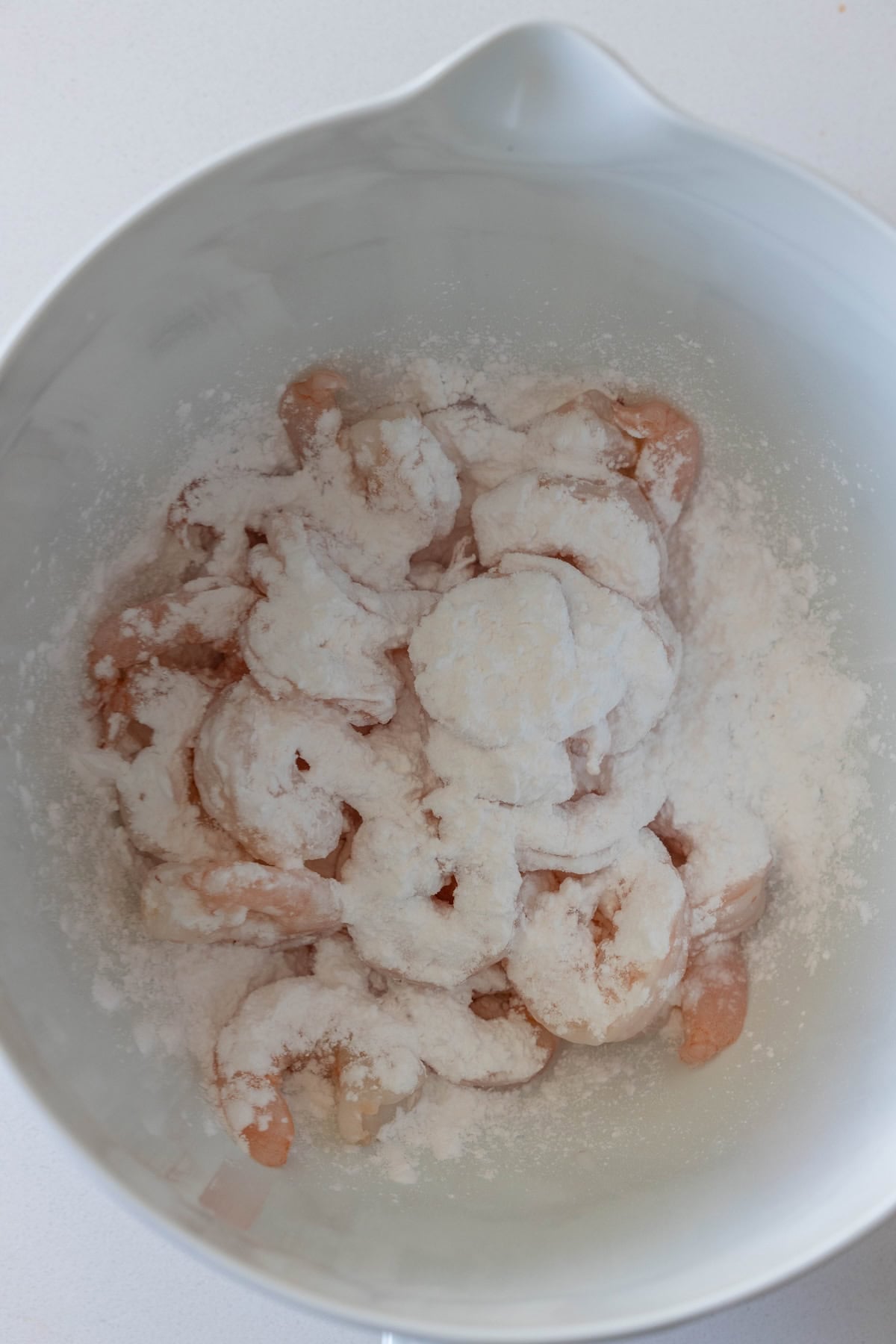 Raw shrimp coated with white cornstarch in a large white mixing bowl on a white surface, perfect for making salt and pepper shrimp.