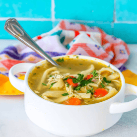 A bowl of chicken noodle soup with vegetables, garnished with parsley, served with a spoon in it. This semi-homemade chicken noodle soup recipe sits perfectly next to a colorful checkered towel in the background.