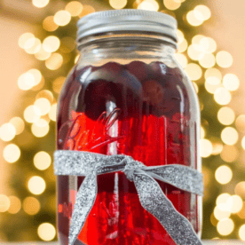 A glass mason jar filled with Cranberry Moonshine is tied with a silver ribbon, set in front of blurred yellow holiday lights.