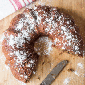 A partially sliced Date Nut Bread bundt cake, dusted with powdered sugar, sits on a wooden board next to a knife and a red-striped kitchen towel.