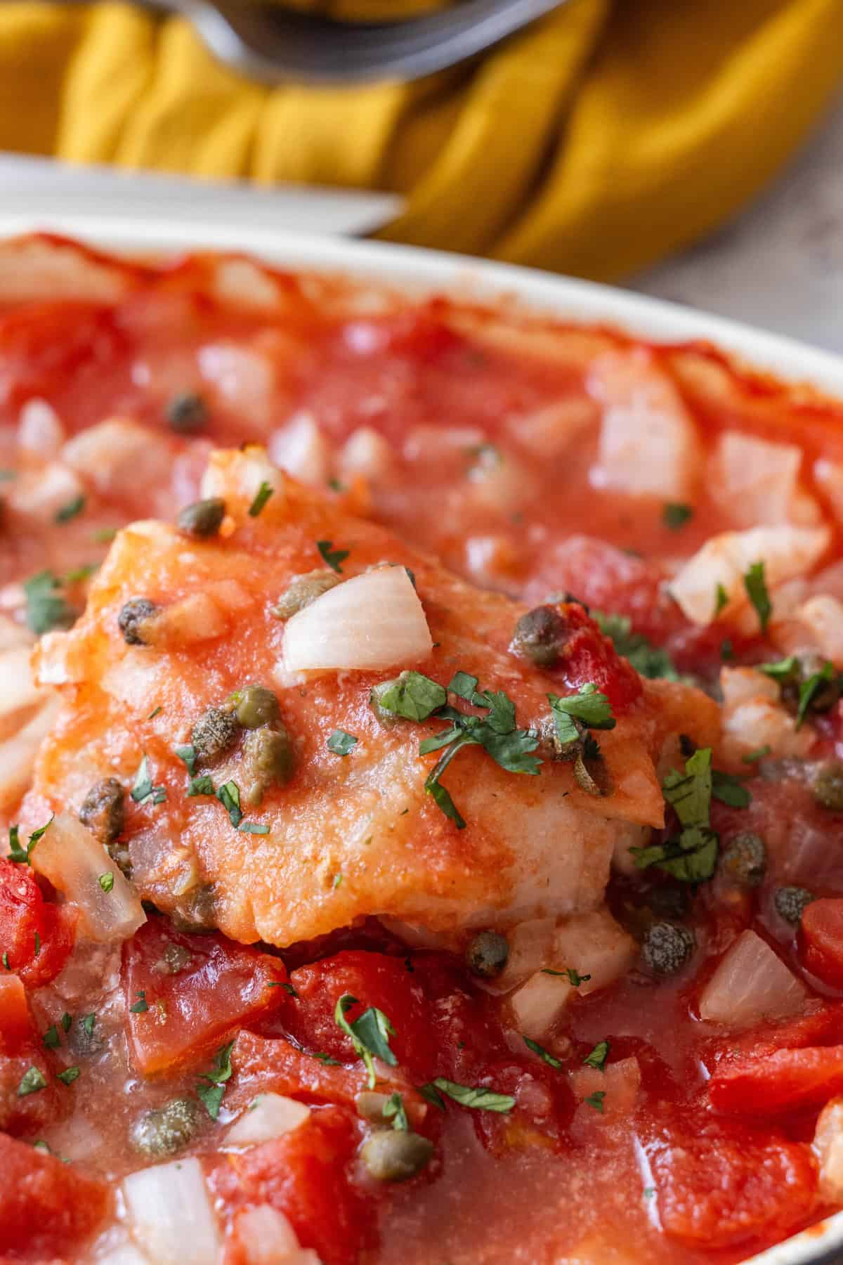 A piece of white fish topped with tomato sauce, diced onions, capers, and chopped herbs on a white plate—an easy baked fish inspired by a Tuscan baked fish recipe.