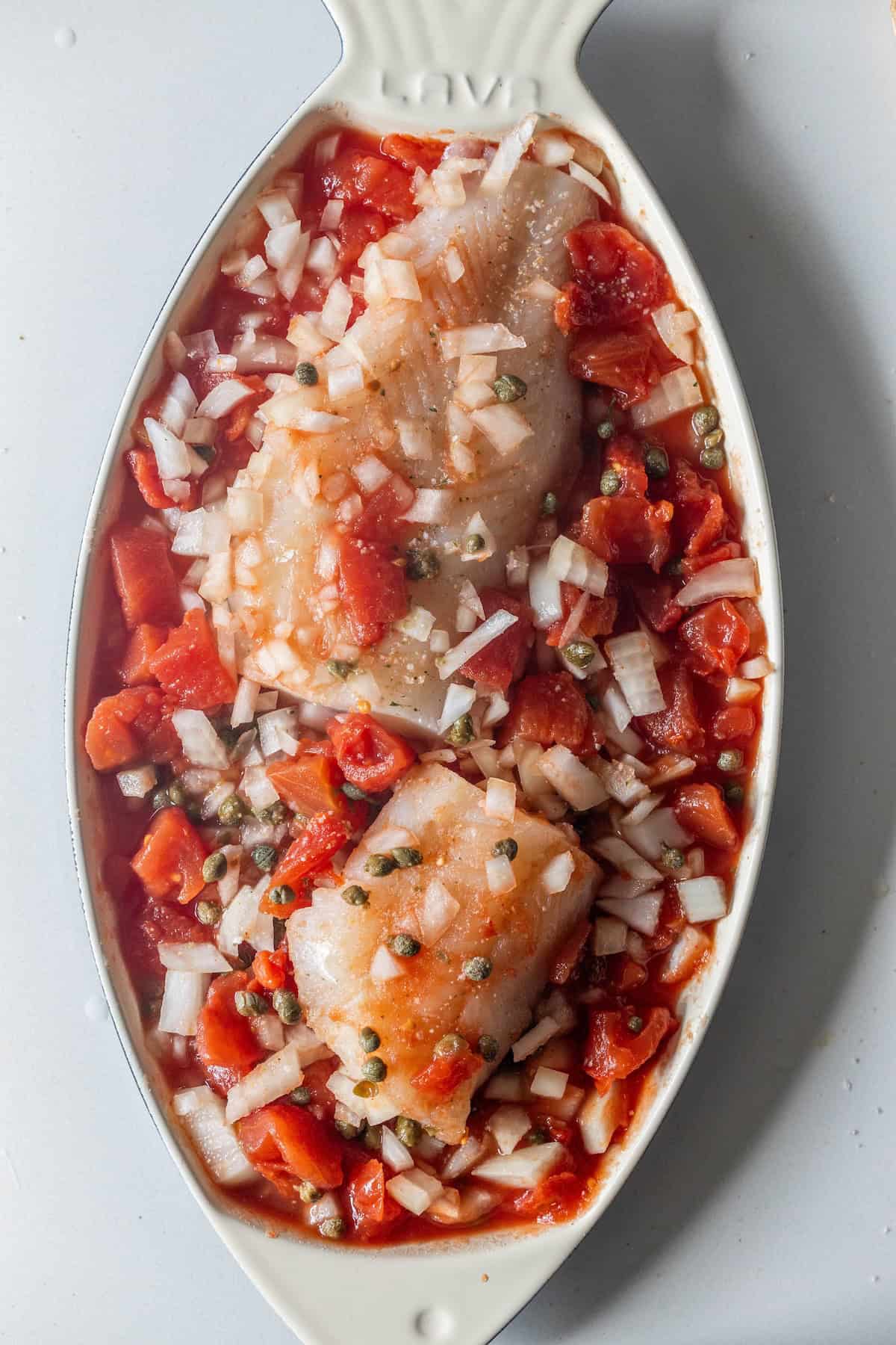 Raw fish fillets topped with chopped onions, diced tomatoes, and capers in a white oval dish, ready for baking—a perfect start for an easy baked fish or Tuscan baked fish recipe.