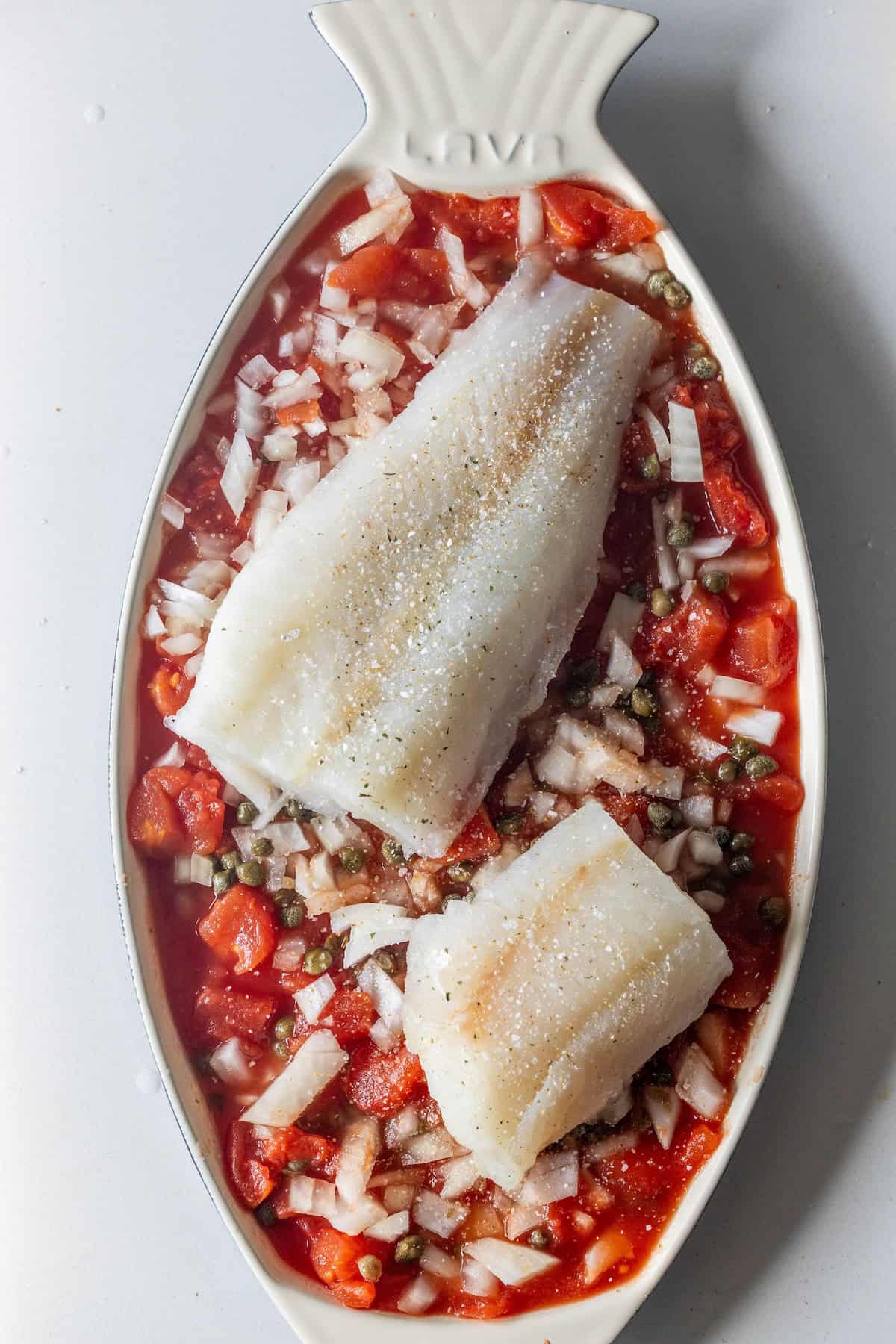 Two raw white fish fillets rest on chopped tomatoes, onions, and capers in a fish-shaped baking dish—an easy baked fish inspired by One Pot 5 Ingredient Tuscan Baked Fish.