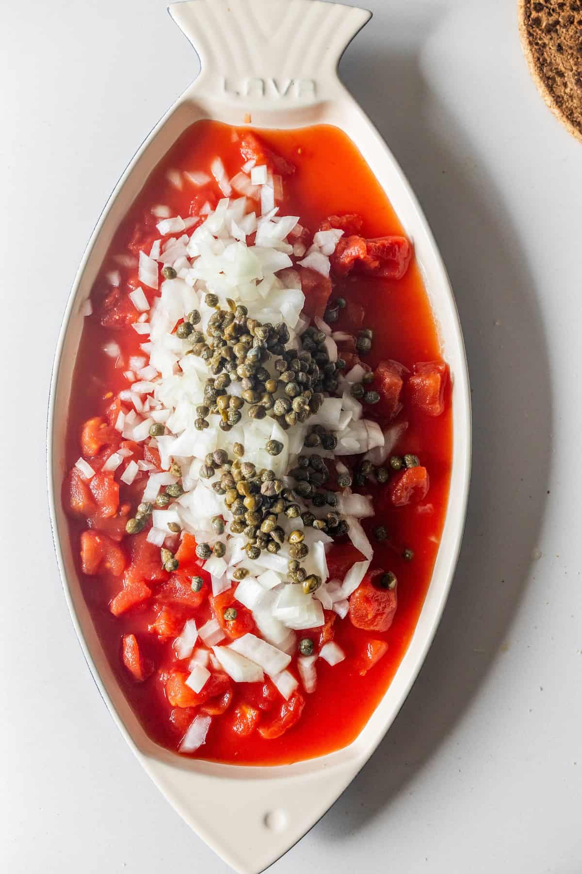 A fish-shaped dish filled with chopped tomatoes, diced onions, and capers in a rich tomato sauce—perfect for those craving a simple One Pot 5 Ingredient Tuscan Baked Fish.
