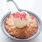 A baked dessert of brown butter blondies in a round pan topped with a scoop of vanilla ice cream, pink and red sprinkles, and heart-shaped candies.