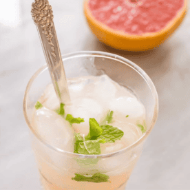 A refreshing pink gimlet mocktail features a glass of iced grapefruit juice adorned with mint leaves, accompanied by a metal spoon and a juicy grapefruit half in the background.