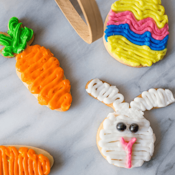 Colorful, easy Easter-themed sugar cookies decorated to resemble a carrot, an easter egg, and a bunny face on a marble surface.