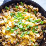 A cast iron skillet sizzles with cheesy veggie home fries, a delightful potato dish crowned with chopped green onions.