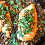 Two cooked chicken breasts garnished with chopped cilantro and diced vegetables in a rich mushroom and onion sauce, served in a skillet.