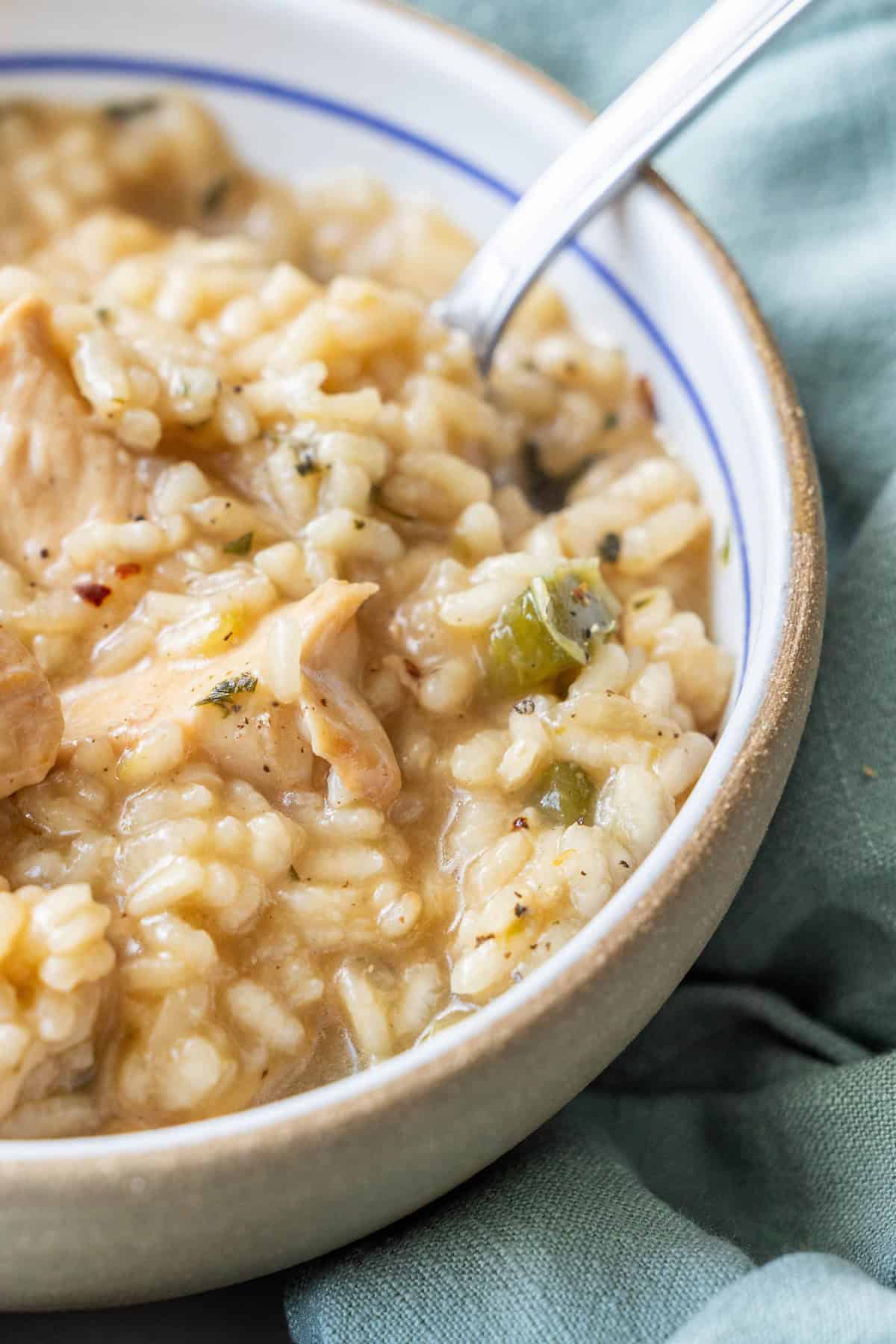 A close-up of a bowl of creamy risotto with chicken pieces, garnished with herbs, and a metal spoon resting in the dish showcases this easy risotto recipe perfectly.