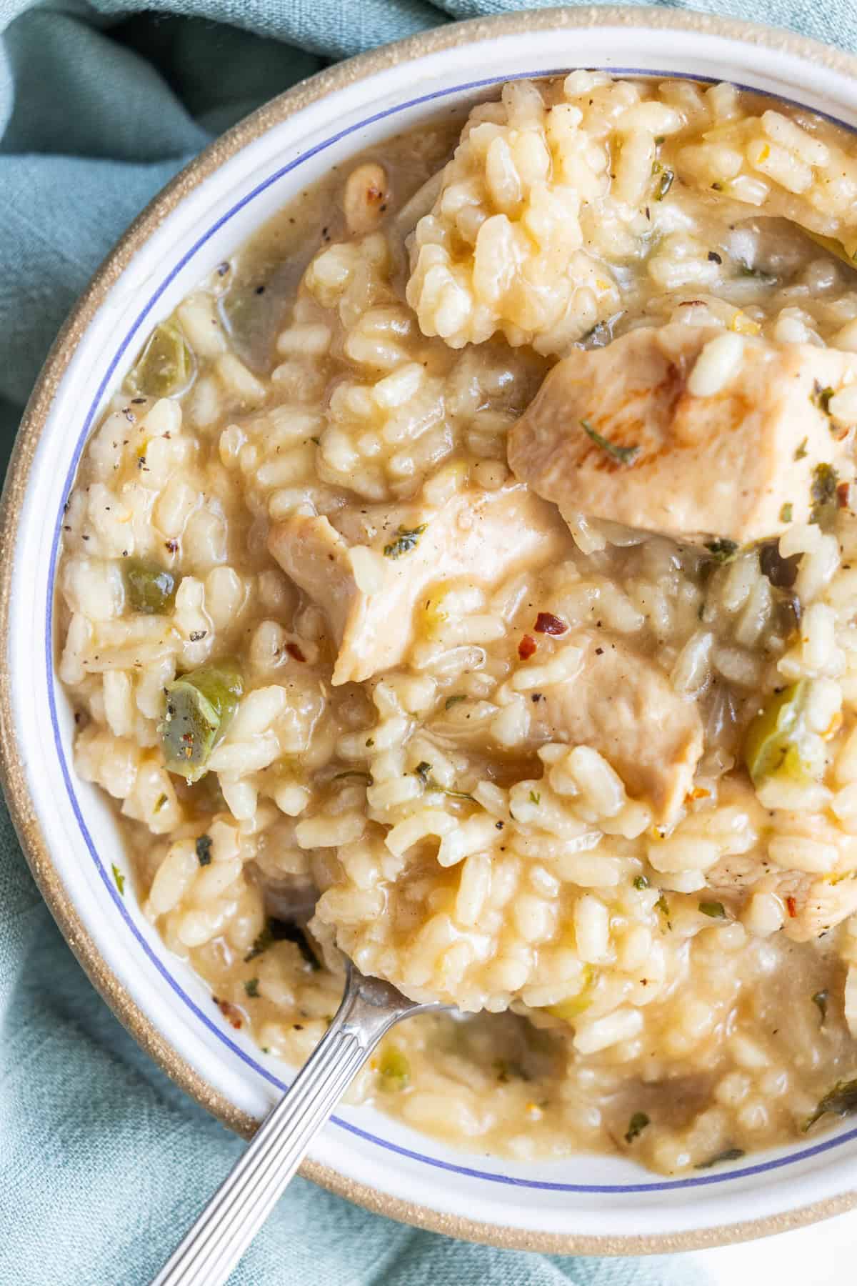 A close-up of a bowl of creamy one pot chicken risotto, featuring tender chunks of chicken, rice, and green vegetables, with a spoon partially dipped in the dish.