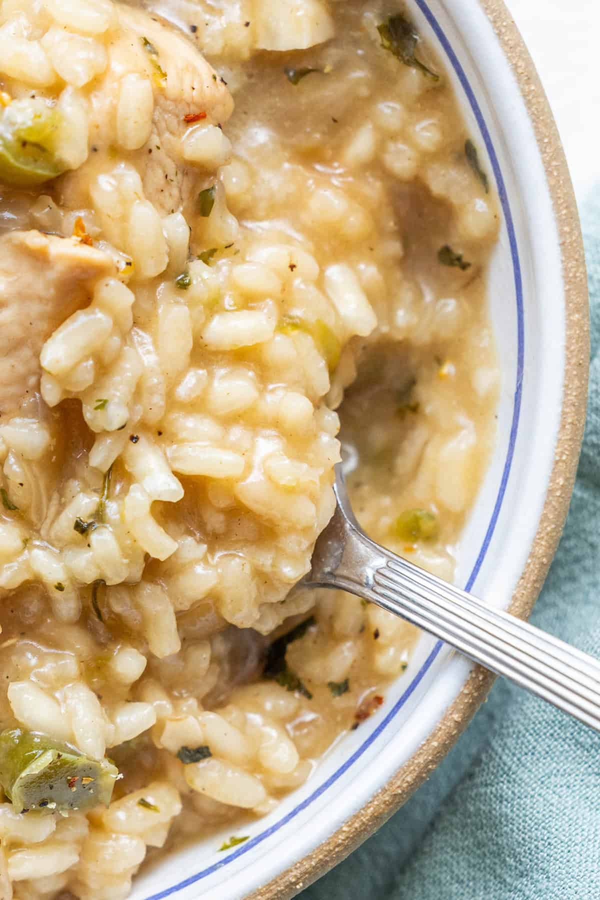 Close-up of a bowl of one pot risotto with chunks of green vegetables and chicken, shown with a silver spoon.