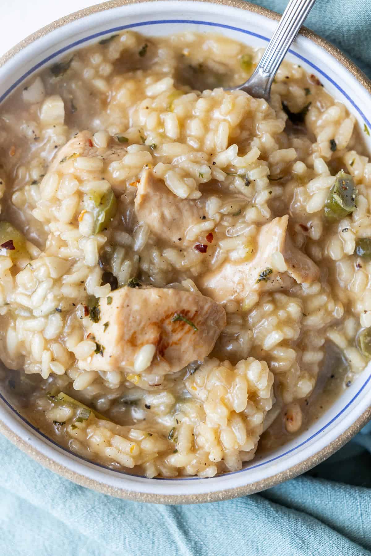 A close-up of a bowl of creamy chicken and rice stew, resembling an easy risotto recipe, with visible chunks of chicken, rice, and bits of vegetables, and a spoon resting in the bowl.