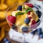 A glass dish filled with fresh raspberries, blackberries, and blueberries is topped with silky Berries and Creme Anglaise sauce and finished with a mint sprig.