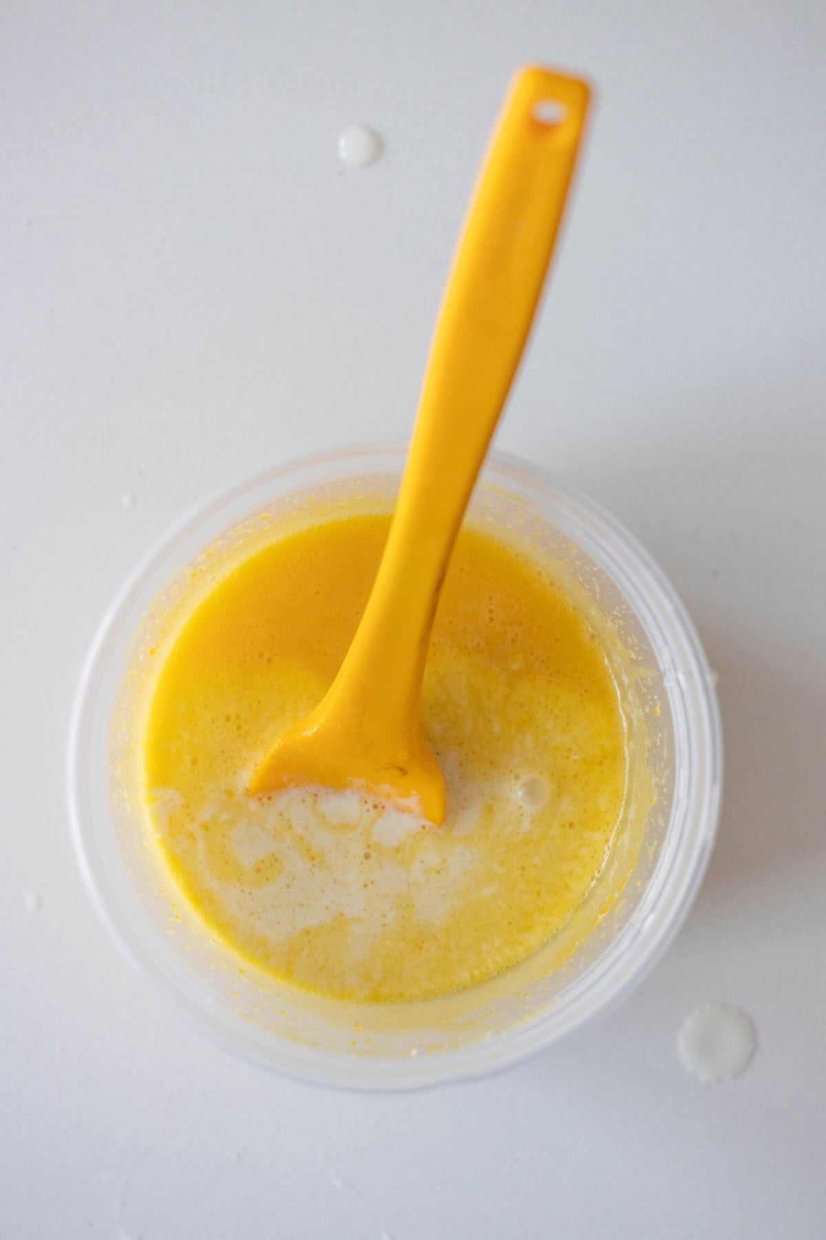 A yellow spoon in a clear plastic container with beaten eggs and a touch of Berries and Creme Anglaise on a white surface, seen from above.