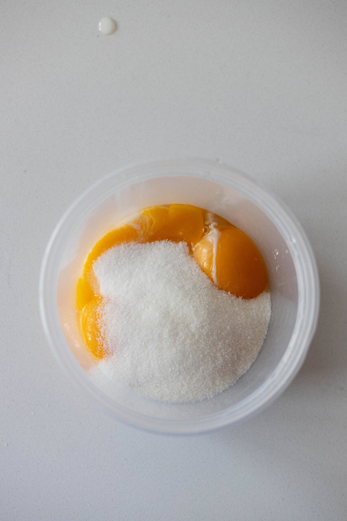 A clear container holding several egg yolks and a pile of granulated white sugar on a light-colored surface, ready to be transformed into a luscious Berries and Creme Anglaise.