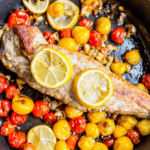 A cooked piece of One Pan Tuscan Pork Loin garnished with lemon slices surrounded by yellow and red cherry tomatoes in a pan.