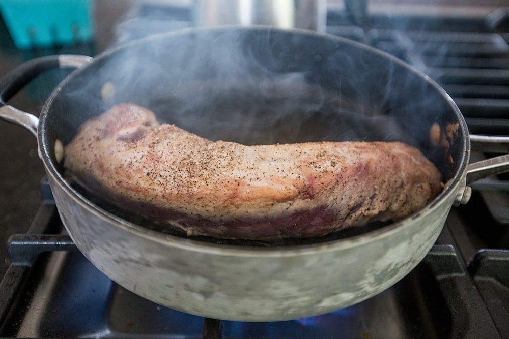 hot pan on stovetop with pork tenderloin searing in it. 