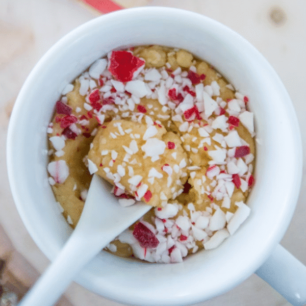 A white mug filled with Microwave Peppermint Cookie Dough Fudge for One, topped with crushed red and white peppermint candy, with a spoon inside the mug.