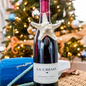 A bottle of wine sits on a wicker basket in front of a beach themed christmas tree.