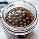 A jar filled with dark brown tapioca pearls, resembling chocolate drops, submerged in liquid, viewed from above on a light surface.