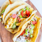 Three spicy egg and chorizo breakfast tacos with scrambled eggs, tomatoes, green onions, and sauce in tortillas on a wooden board.