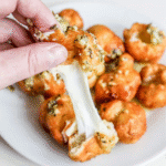 A hand pulls apart a Fried Garlic Cheese Bomb, stretching melted cheese, with more golden bread bites on a white plate in the background.
