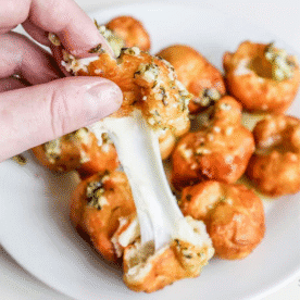 A hand pulls apart a Fried Garlic Cheese Bomb, stretching melted cheese, with more golden bread bites on a white plate in the background.