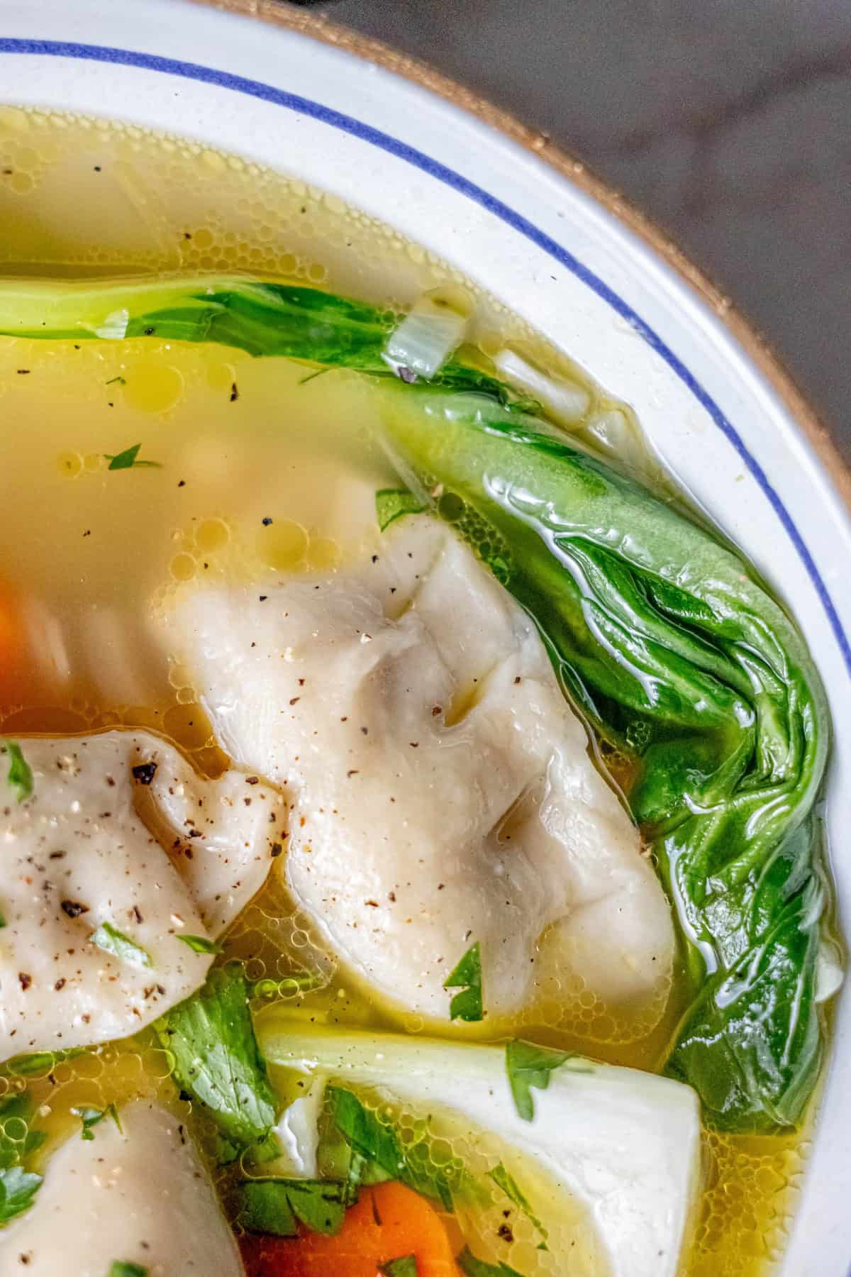 Close-up of a quick 15-minute potsticker soup featuring dumplings, carrots, bok choy, and herbs in a clear broth served in a white bowl with a blue rim.