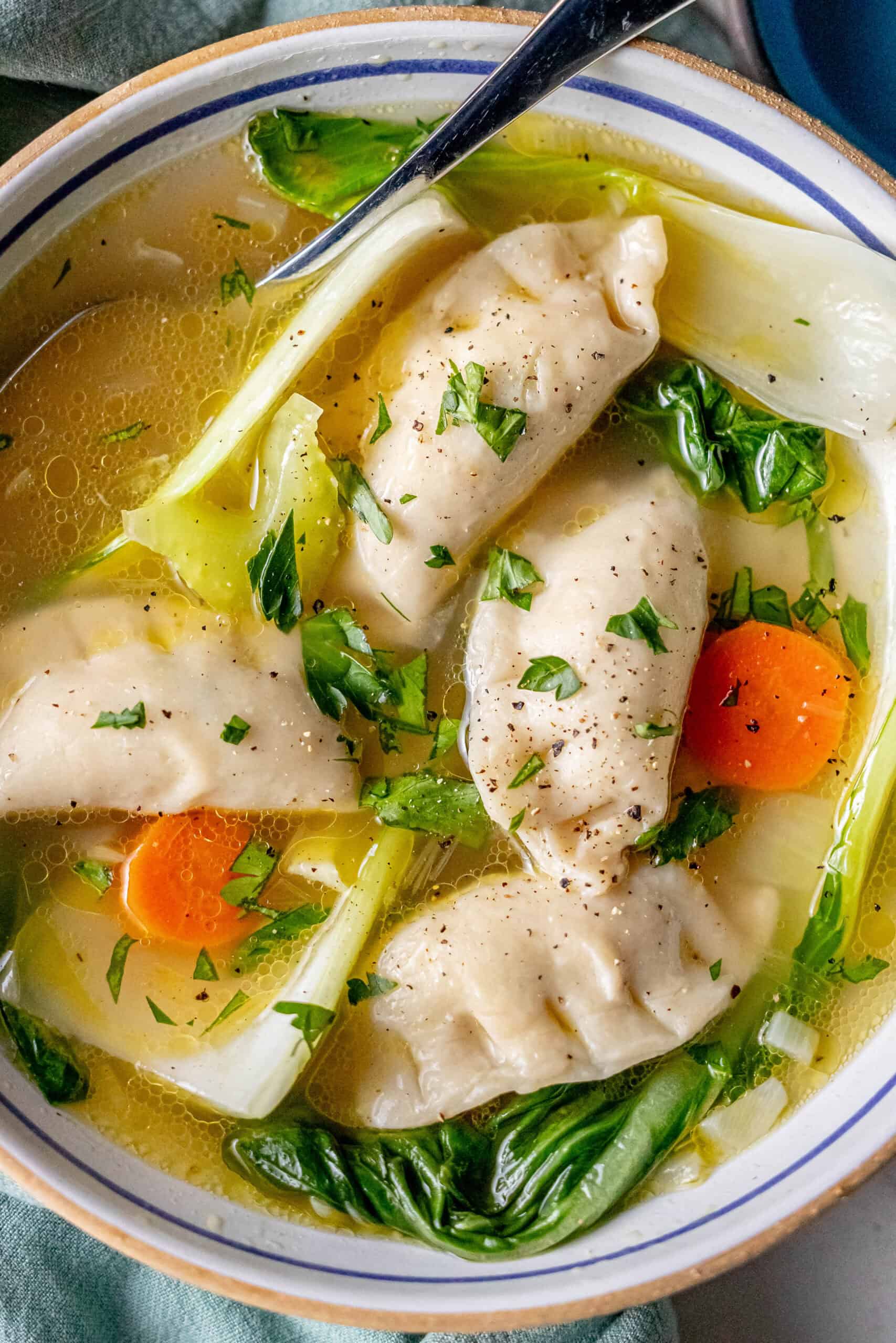 A bowl of 15-minute potsticker soup holds three dumplings amid leafy greens, carrot slices, and savory broth, garnished with chopped herbs. A spoon rests in the bowl.