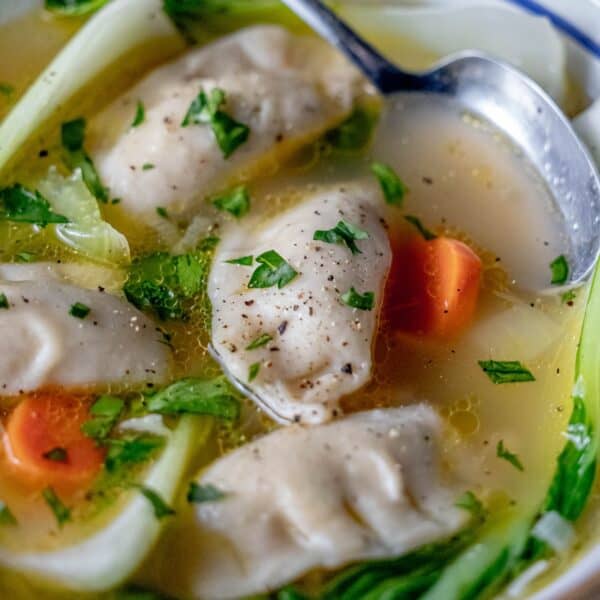 A bowl of 15-minute potsticker soup brimming with plump dumplings, carrots, and greens, garnished with herbs and freshly ground pepper. A spoon waits invitingly on the side.