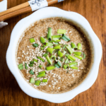 A bowl of miso soup enhanced with shoyu, topped with chopped green onions and sesame seeds, sits on a wooden table, flanked by chopsticks.