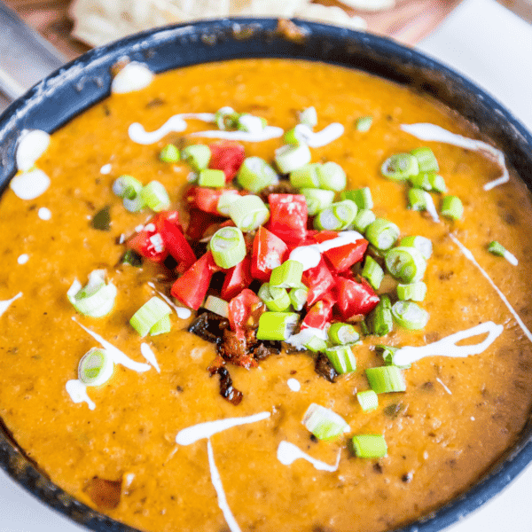 A bowl of spicy soup garnished with diced tomatoes, green onions, and a drizzle of cream.